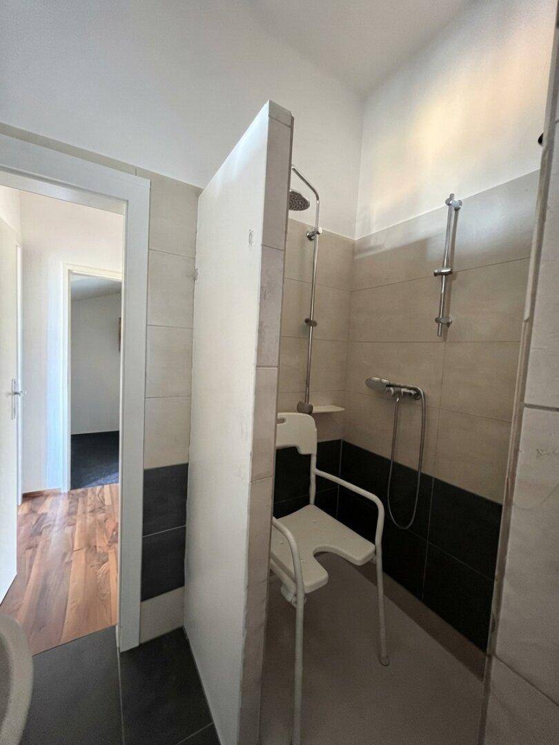 Barrierefreie Dusche mit Fliesenwänden und Duschstuhl für zusätzlichen Komfort.
