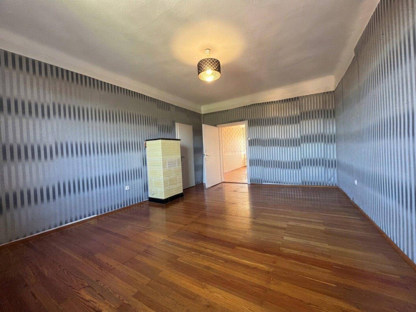 Geräumiges Zimmer mit Holzboden und gestreifter Tapete, ein Kachelofen sorgt für traditionellen Charme.