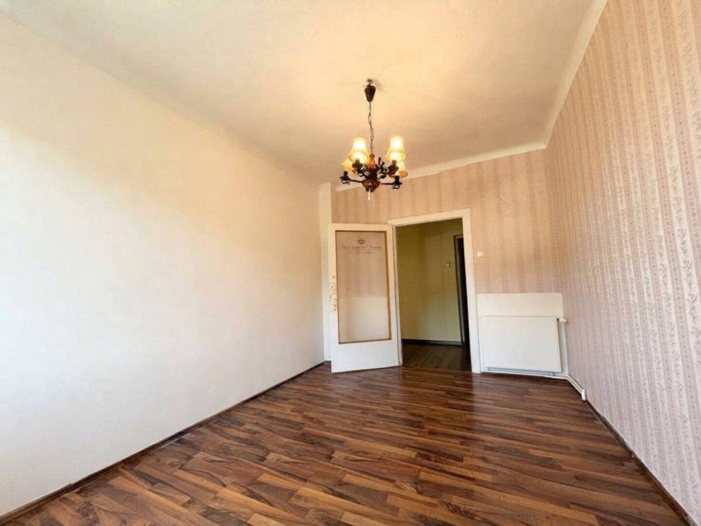 Leeres Zimmer mit Laminatboden und gemusterter Tapete, ein Durchgang führt zu einem angrenzenden Bereich.
