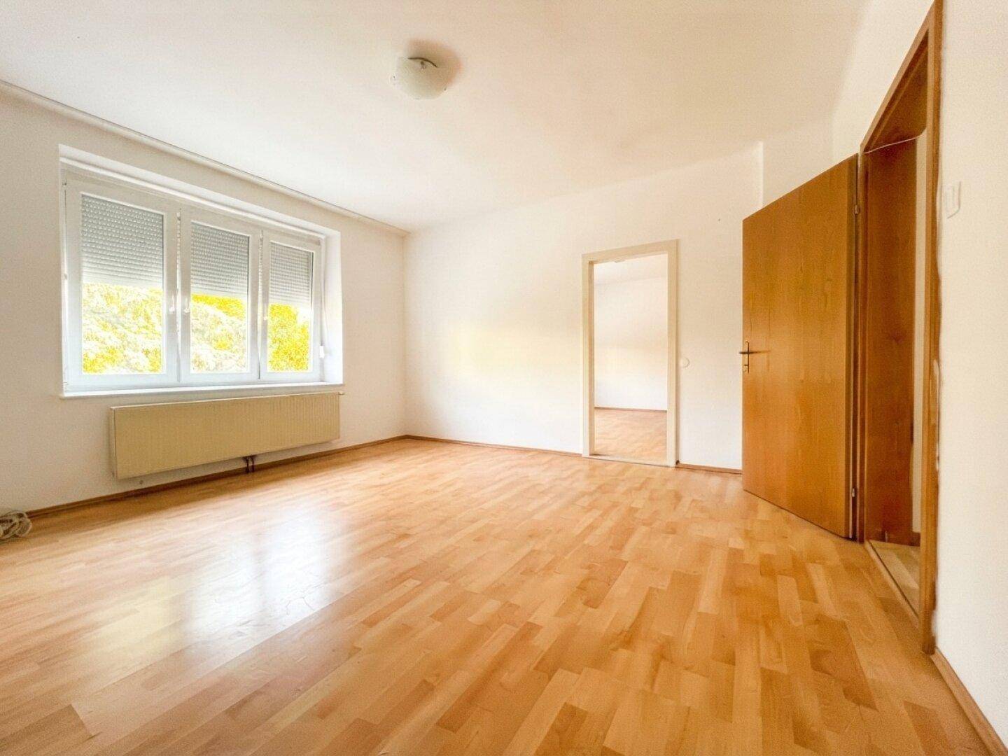 Helles, leeres Zimmer mit Laminatboden und großem Fenster mit Blick ins Grüne.