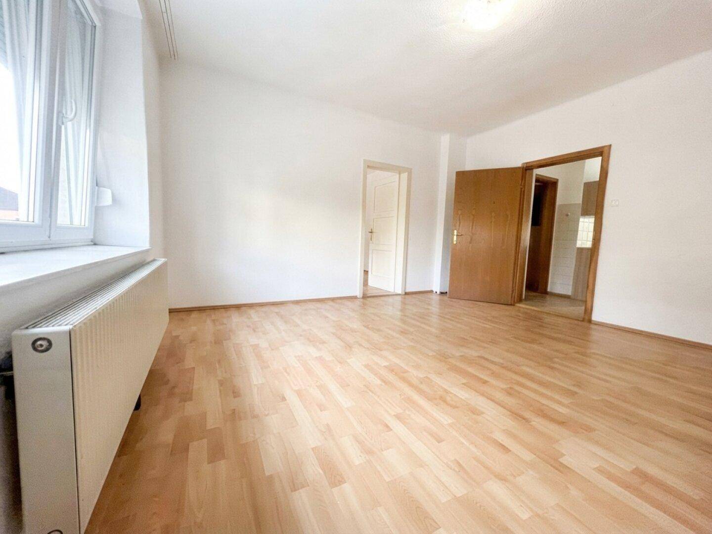 Geräumiges, leeres Zimmer mit Laminatboden und Heizkörper unter dem Fenster.