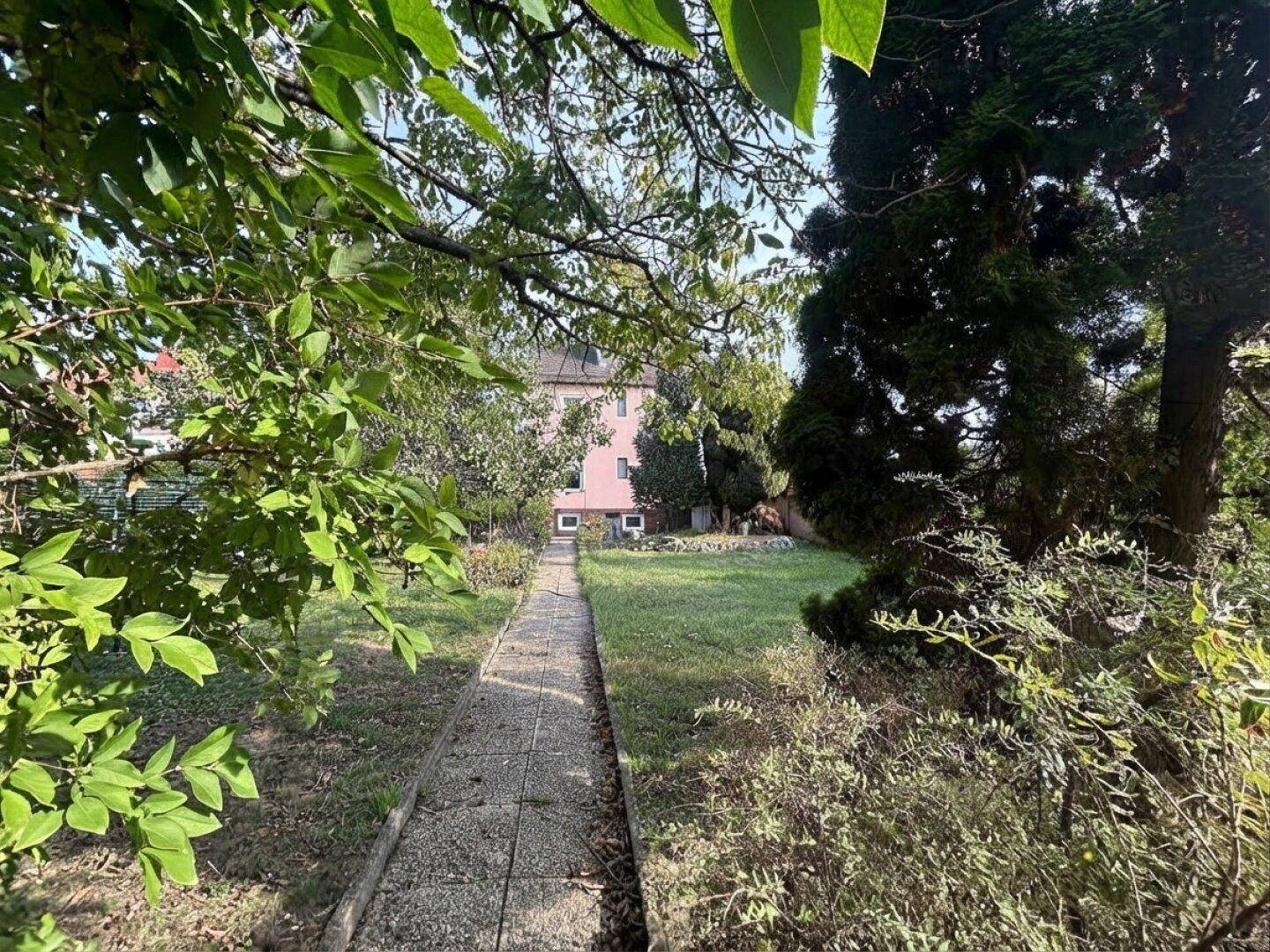 Gartenweg führt durch üppiges Grün zu einem rosafarbenen Gebäude im Hintergrund.
