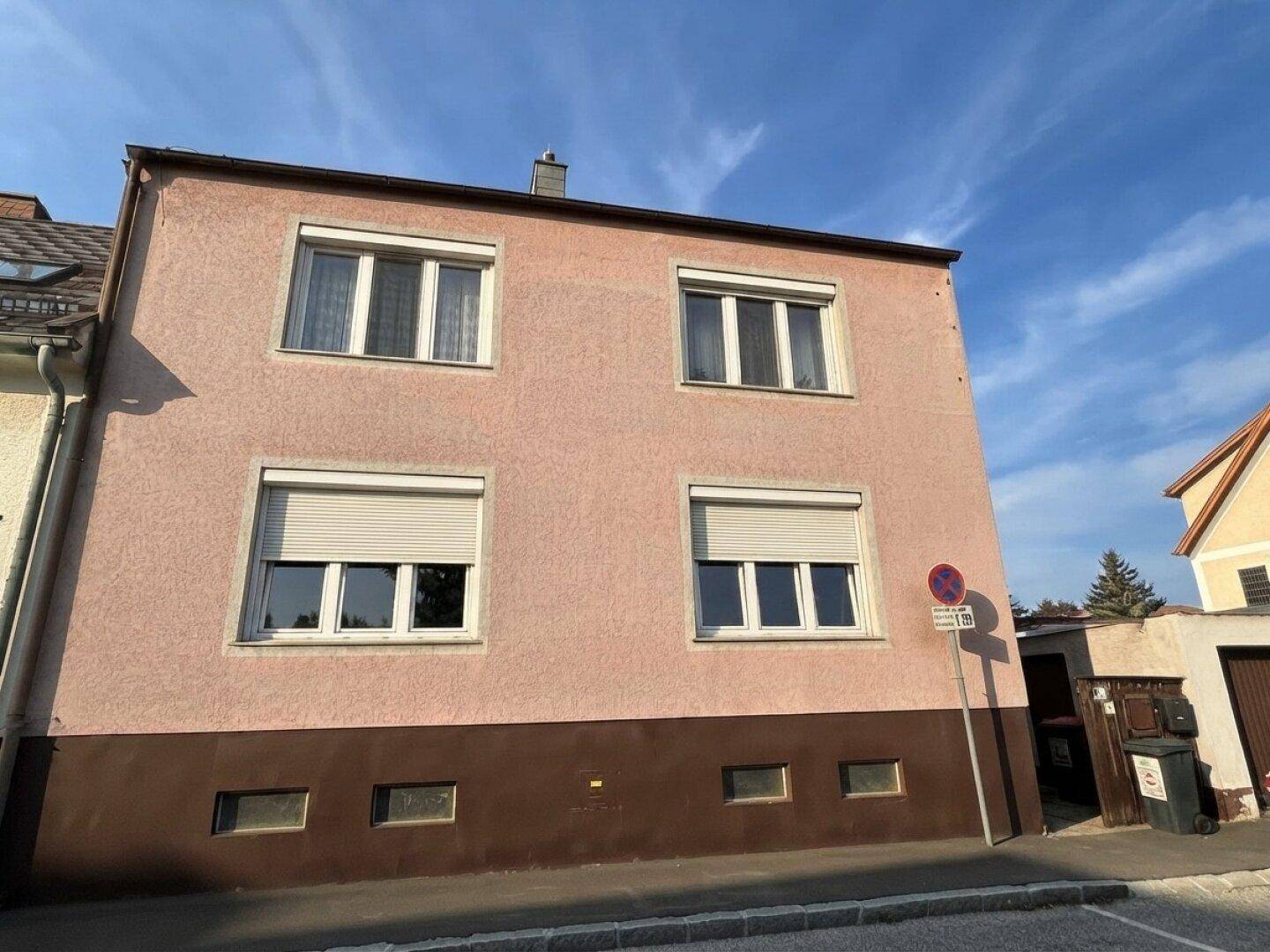 Vorderansicht eines rosafarbenen Wohnhauses mit vier Fenstern und Rollläden zur Straße hin.