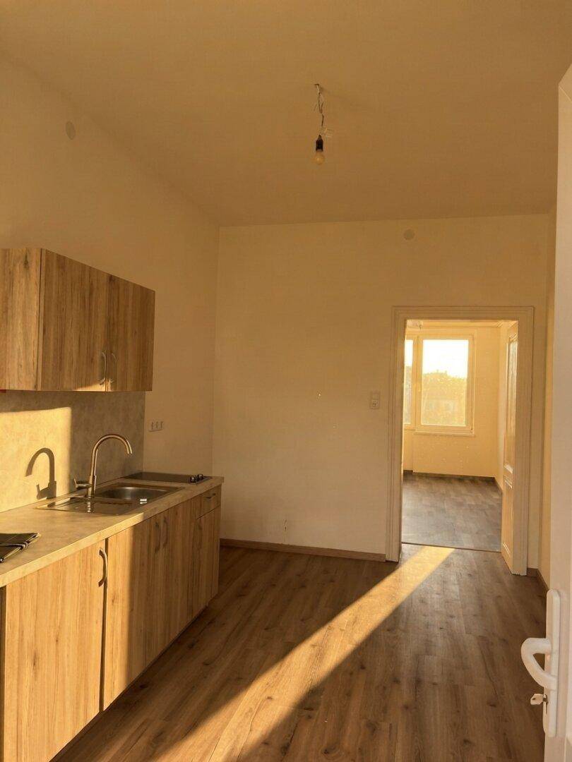 Küchenbereich mit Holzoptik-Boden und einem Durchgang zu einem hellen Nebenraum, durchflutet von Sonnenlicht.