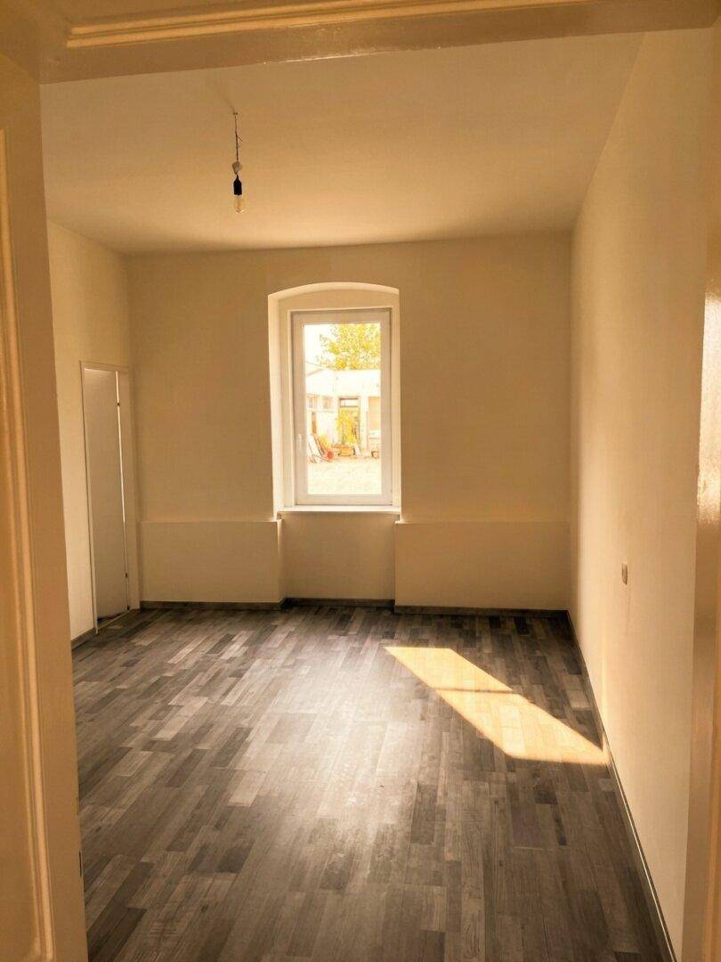 Heller, leerer Raum mit Laminatboden und einem Fenster, das viel natürliches Licht hereinlässt.
