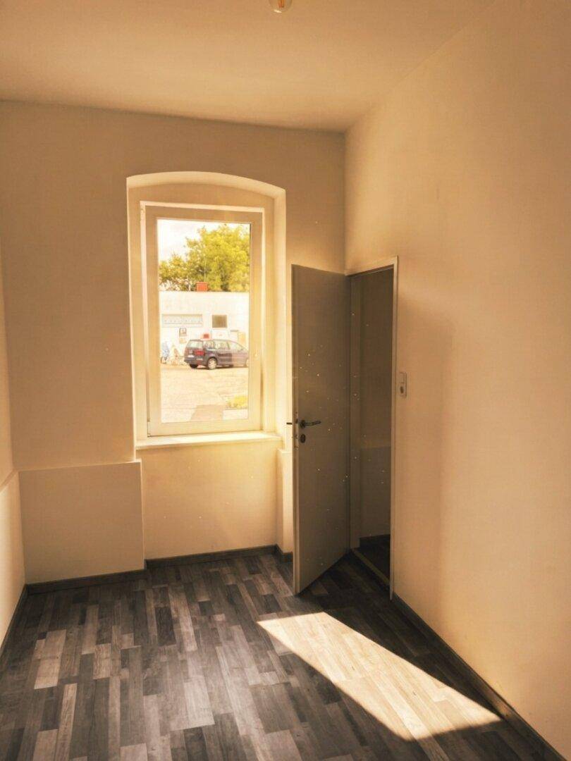 Leeres Zimmer mit Laminatboden und einem Fenster, das den Blick auf die Straße freigibt.