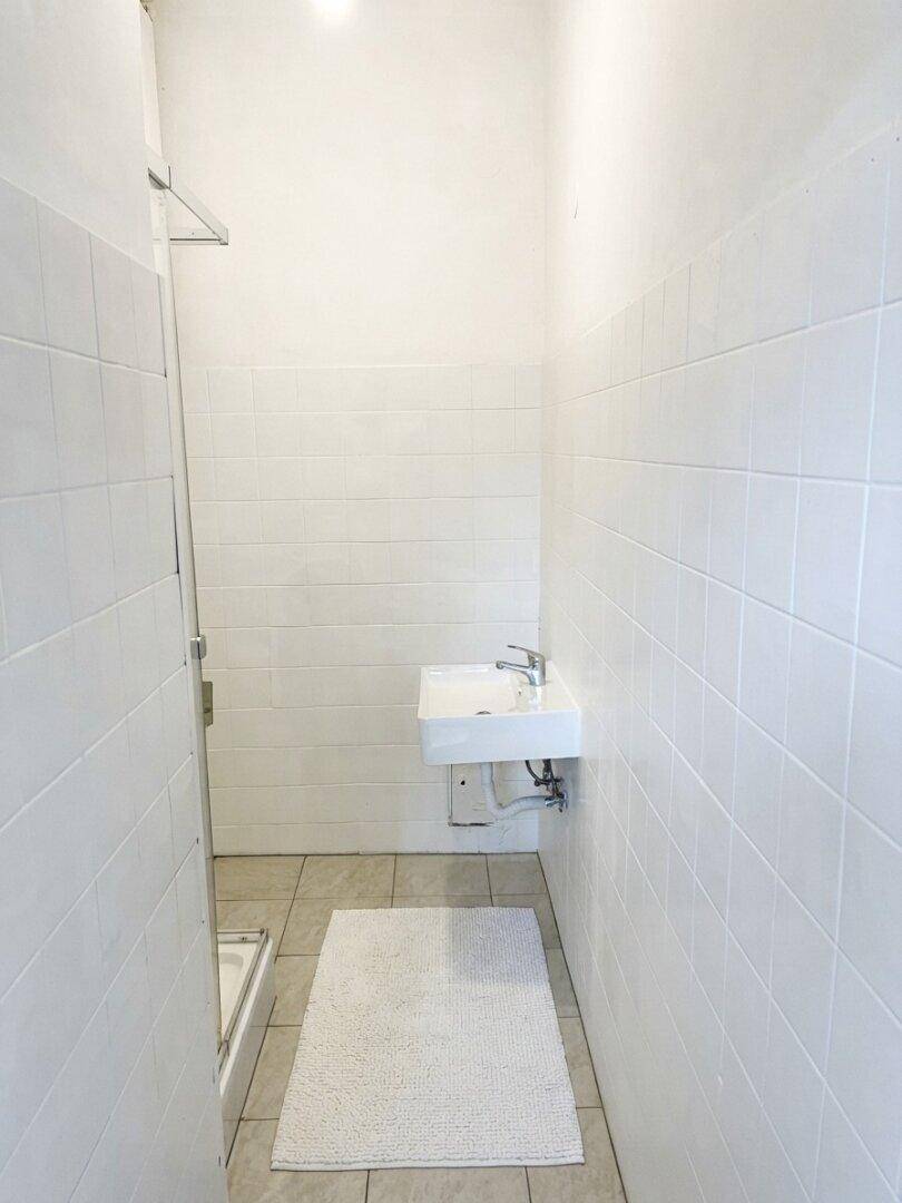 Schlichtes Badezimmer mit weißen Fliesen an Wänden und Boden, ausgestattet mit einem kleinen Waschbecken und Dusche.