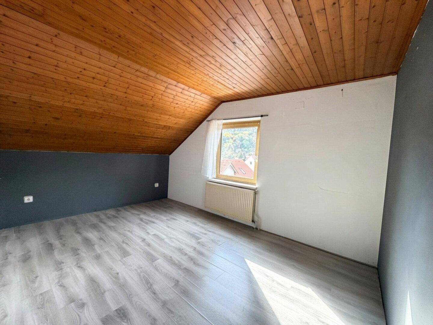 Dachgeschosszimmer mit schräger Decke, Laminatboden und kleinem Fenster, bietet gemütlichen Wohnraum.