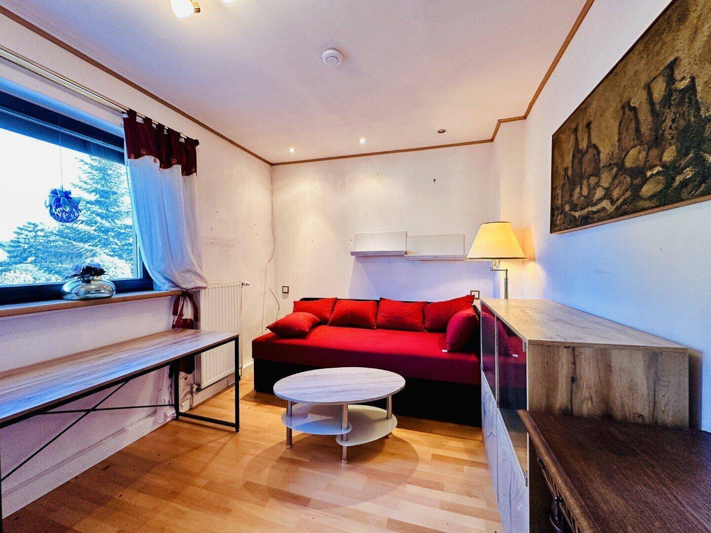 Gemütliches Wohnzimmer mit Holzboden, rotem Sofa und großem Fenster mit Blick ins Grüne.