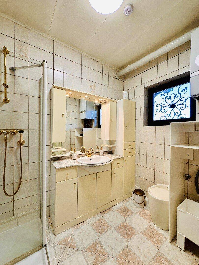Gefliestes Badezimmer mit Dusche, Waschtisch und kleinem Fenster.