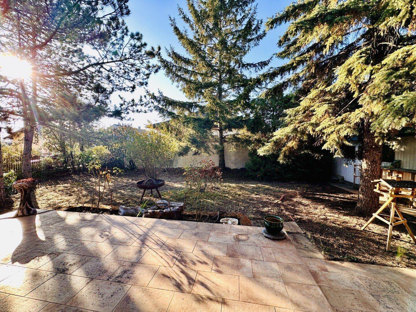 Sonniger Gartenbereich mit Terrasse, Bäumen und Feuerstelle im Hintergrund.