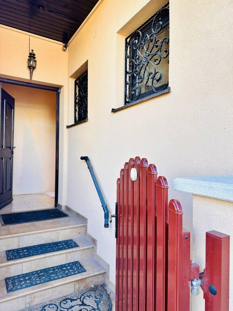 Eingangsbereich eines Hauses mit Steintreppe, schmiedeeisernen Fenstergittern und einem roten Gartentor.