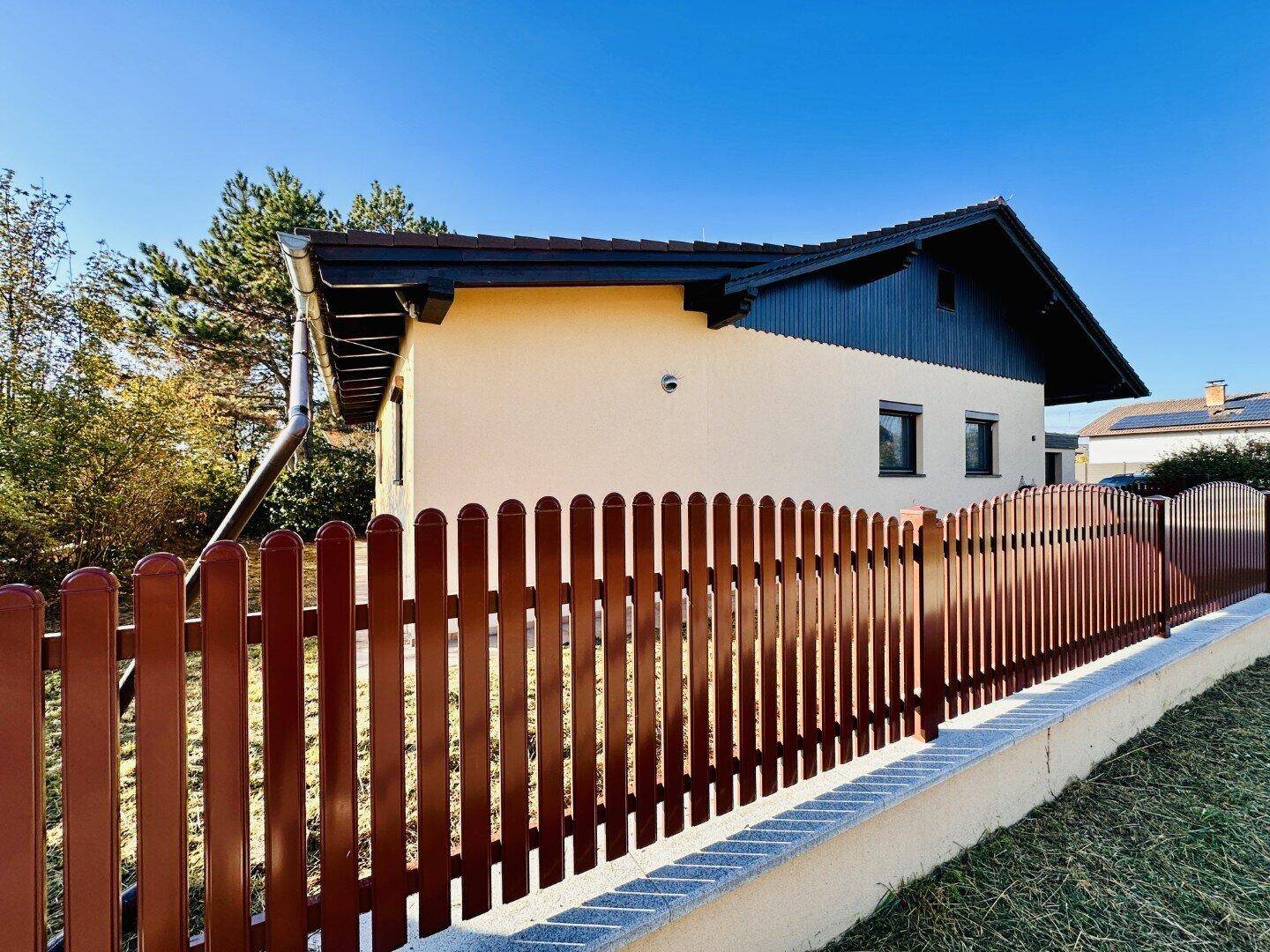 Seitenansicht eines gepflegten Hauses mit beiger Fassade, dunkelblauem Dachgiebel und braunem Lattenzaun.