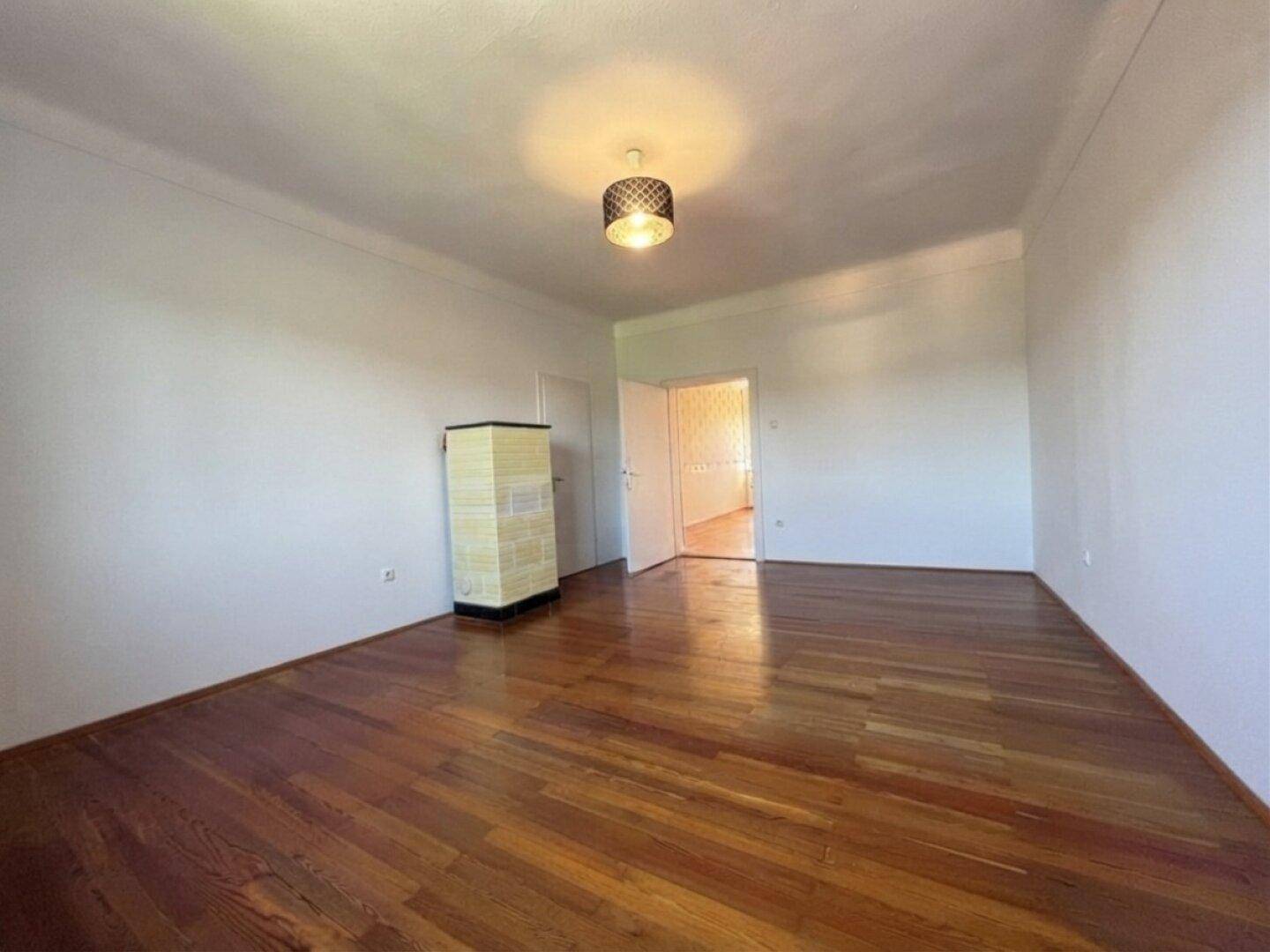 Geräumiges Zimmer mit Holzboden, Kachelofen und Zugang zu weiteren Räumen.