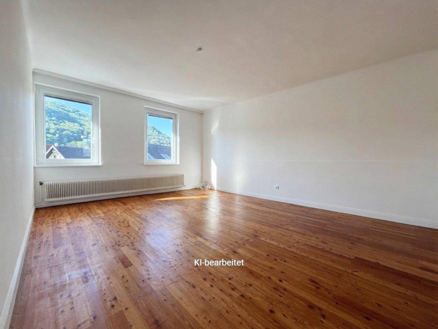 Helles, leeres Zimmer mit Holzboden und zwei Fenstern, die einen Blick ins Grüne bieten.