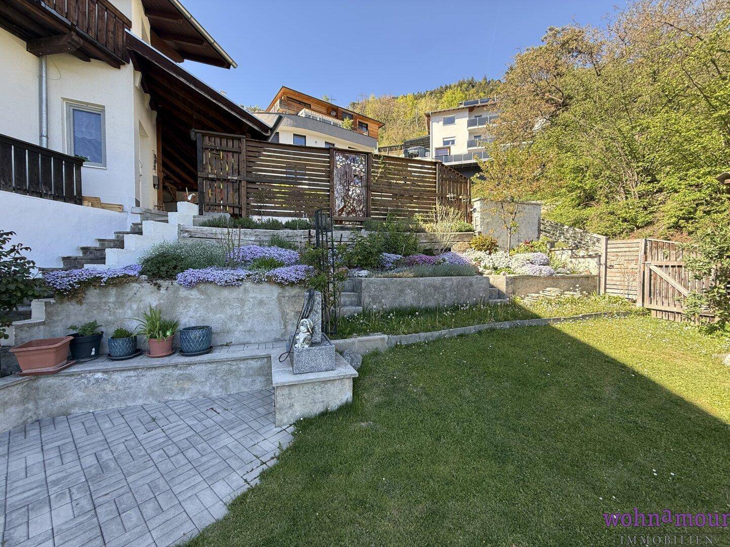 Gestufter Gartenbereich mit Blumenbeeten und gepflasterten Wegen, umgeben von Natur.
