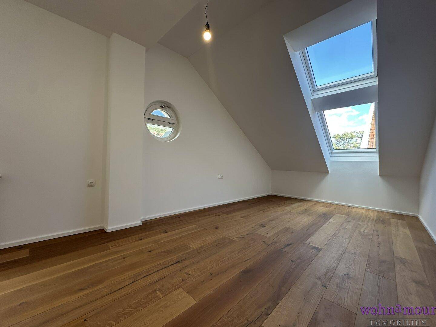 Helles Zimmer im Dachgeschoss mit Holzboden, Dachfenstern und einem runden Fenster.