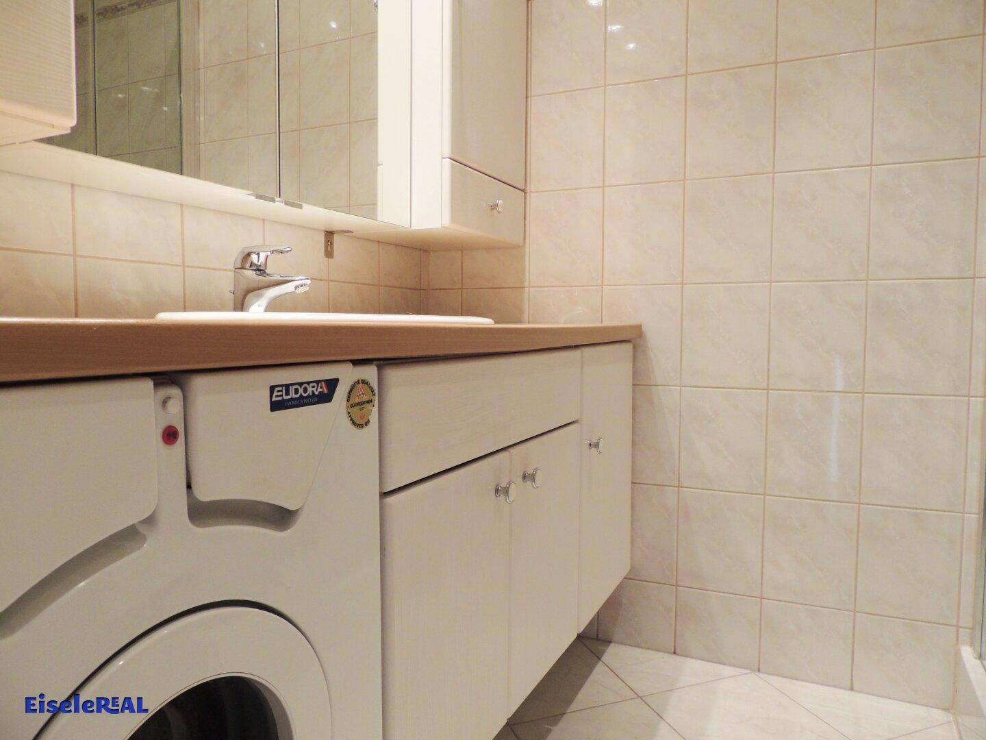 Badezimmer mit Waschbecken, Unterschrank und integrierter Waschmaschine, umgeben von hellen Fliesen.
