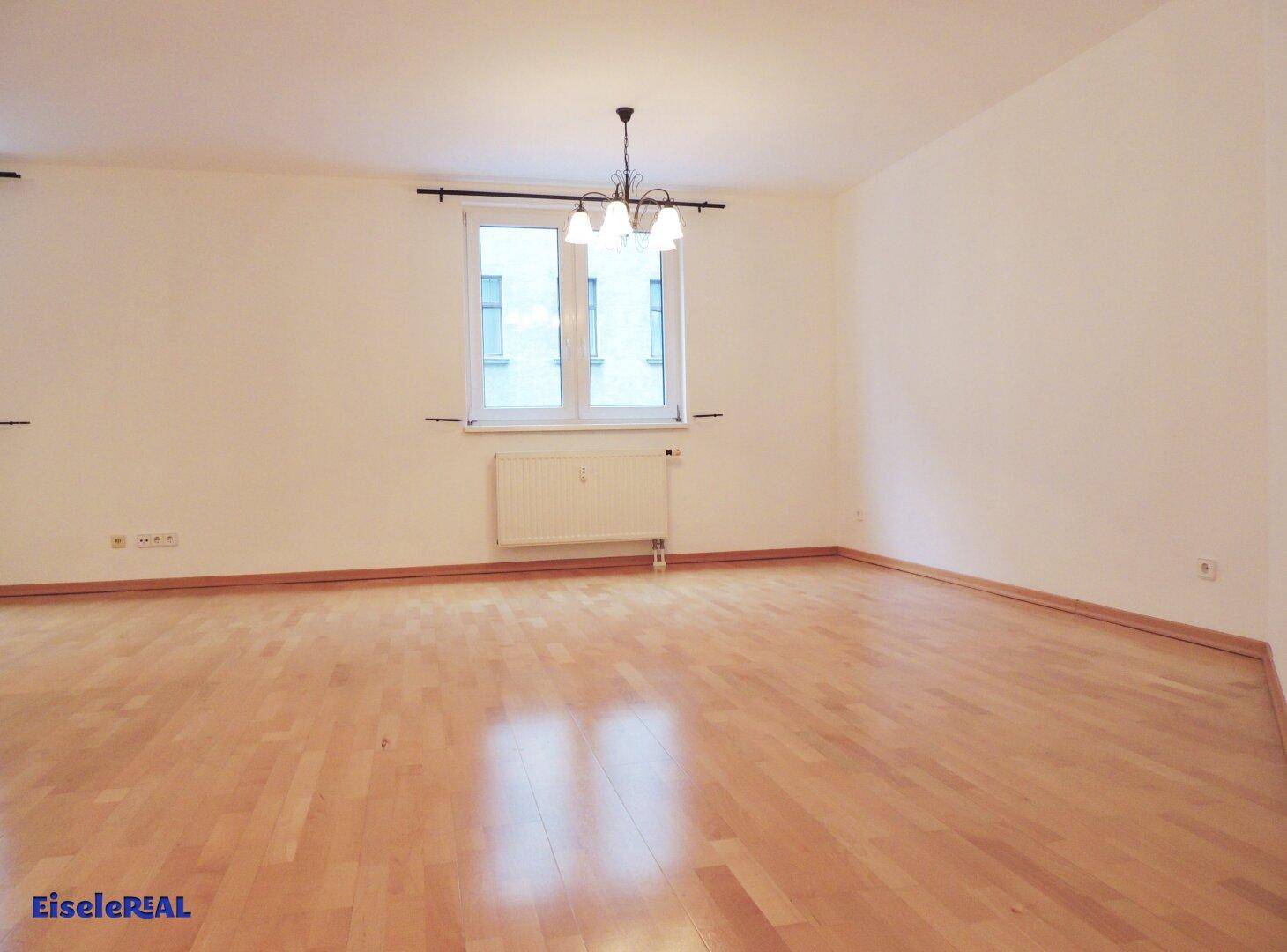 Helles, leeres Wohnzimmer mit Parkettboden und einem Fenster, das viel Tageslicht hereinlässt.