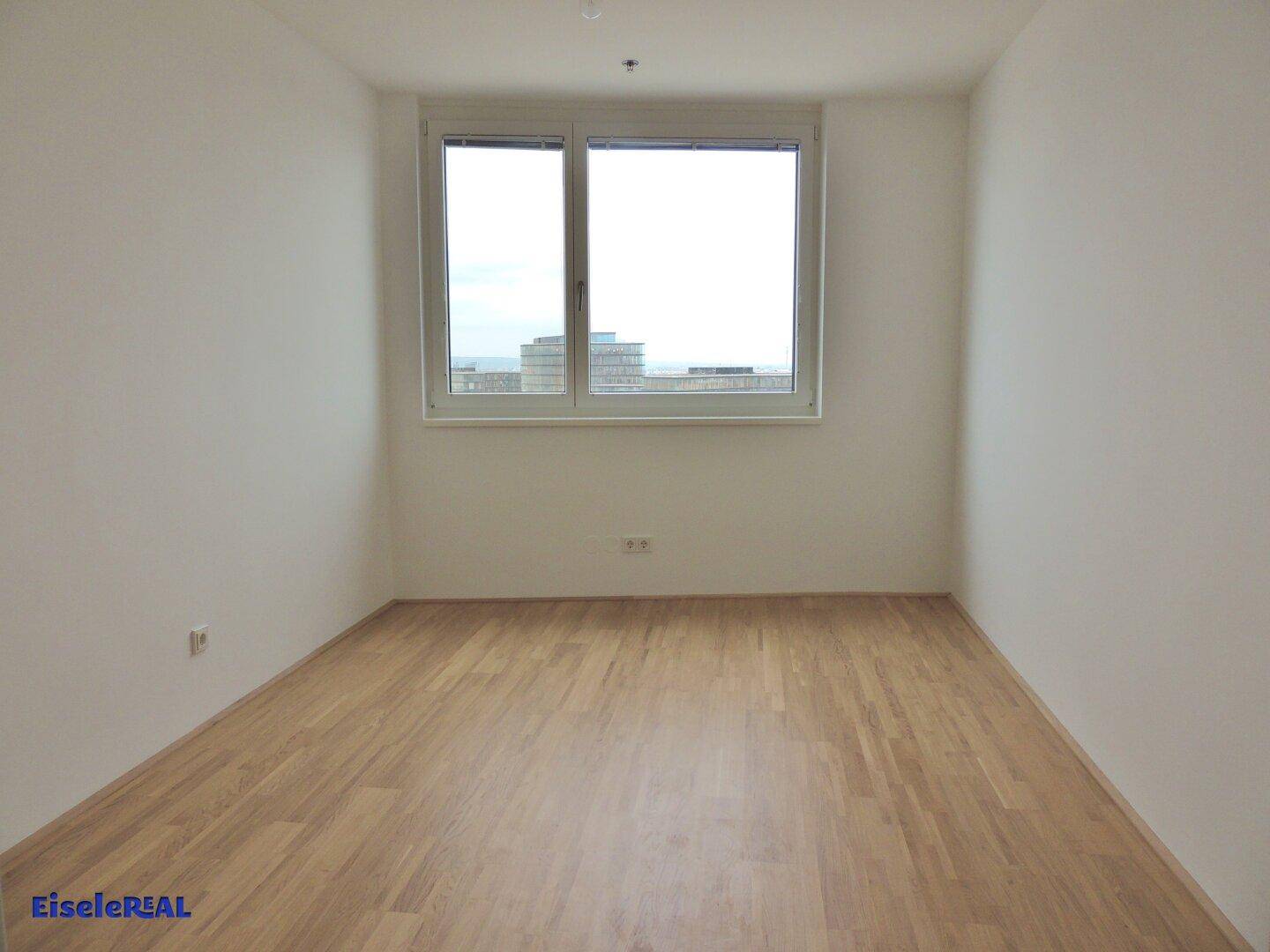 Helles und leeres Zimmer mit Holzboden und einem Fenster, das einen Blick nach draußen ermöglicht.