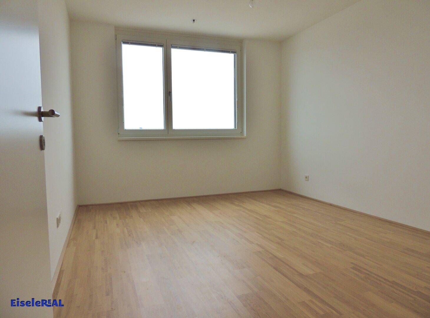 Leeres Schlafzimmer mit Holzboden und einem großen Fenster, das viel Tageslicht bietet.