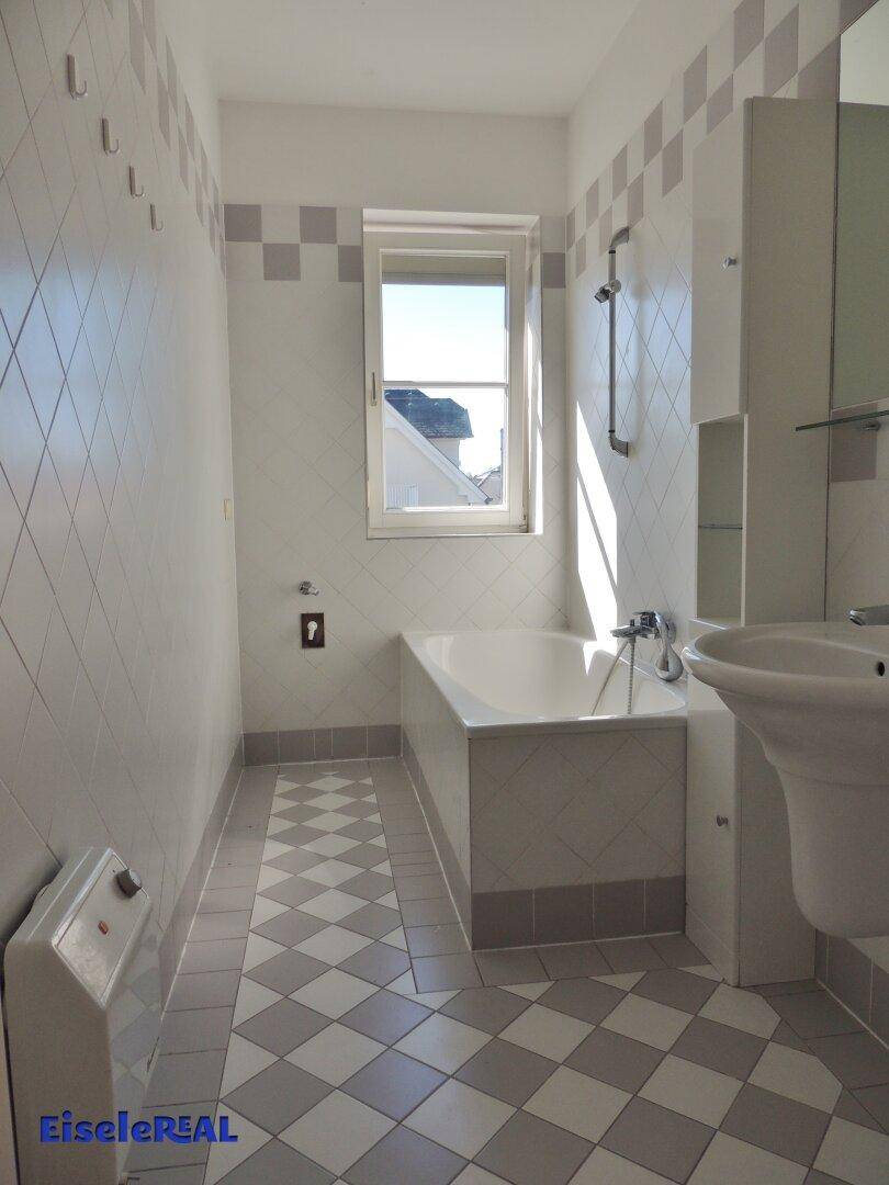 Badezimmer mit Badewanne, Waschbecken und Fenster, gefliest in hellen Tönen.