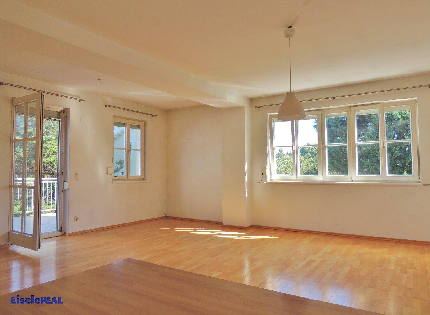 Helles, leeres Wohnzimmer mit Parkettboden und Zugang zu einem Balkon.