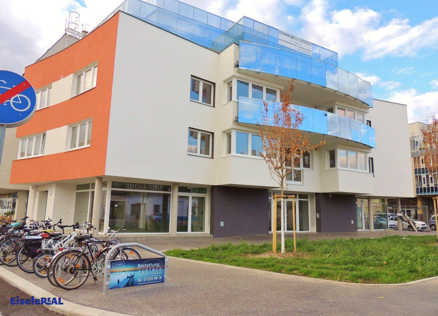 Modernes Gebäude mit weißer und orangefarbener Fassade, blauen Glasbalkonen und Fahrradständern im Vordergrund an einer Straße.
