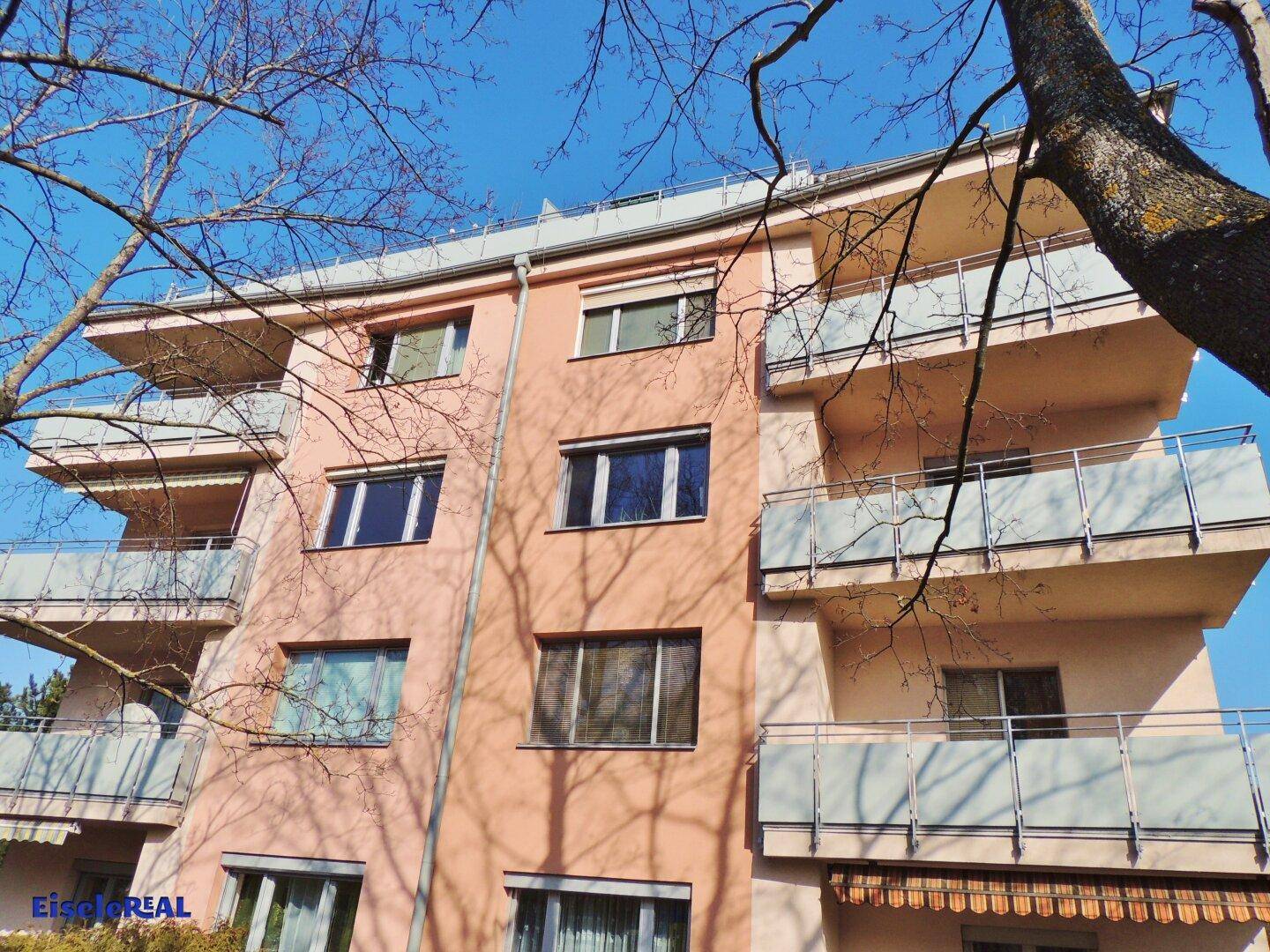 Ansicht des Wohngebäudes mit Balkonen und Fenstern, umgeben von kahlen Bäumen im Frühling.