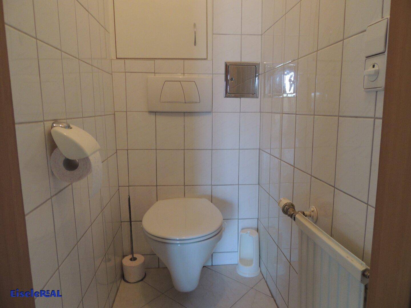 Separates WC mit wandhängender Toilette, Heizkörper und hellen Wand- und Bodenfliesen.