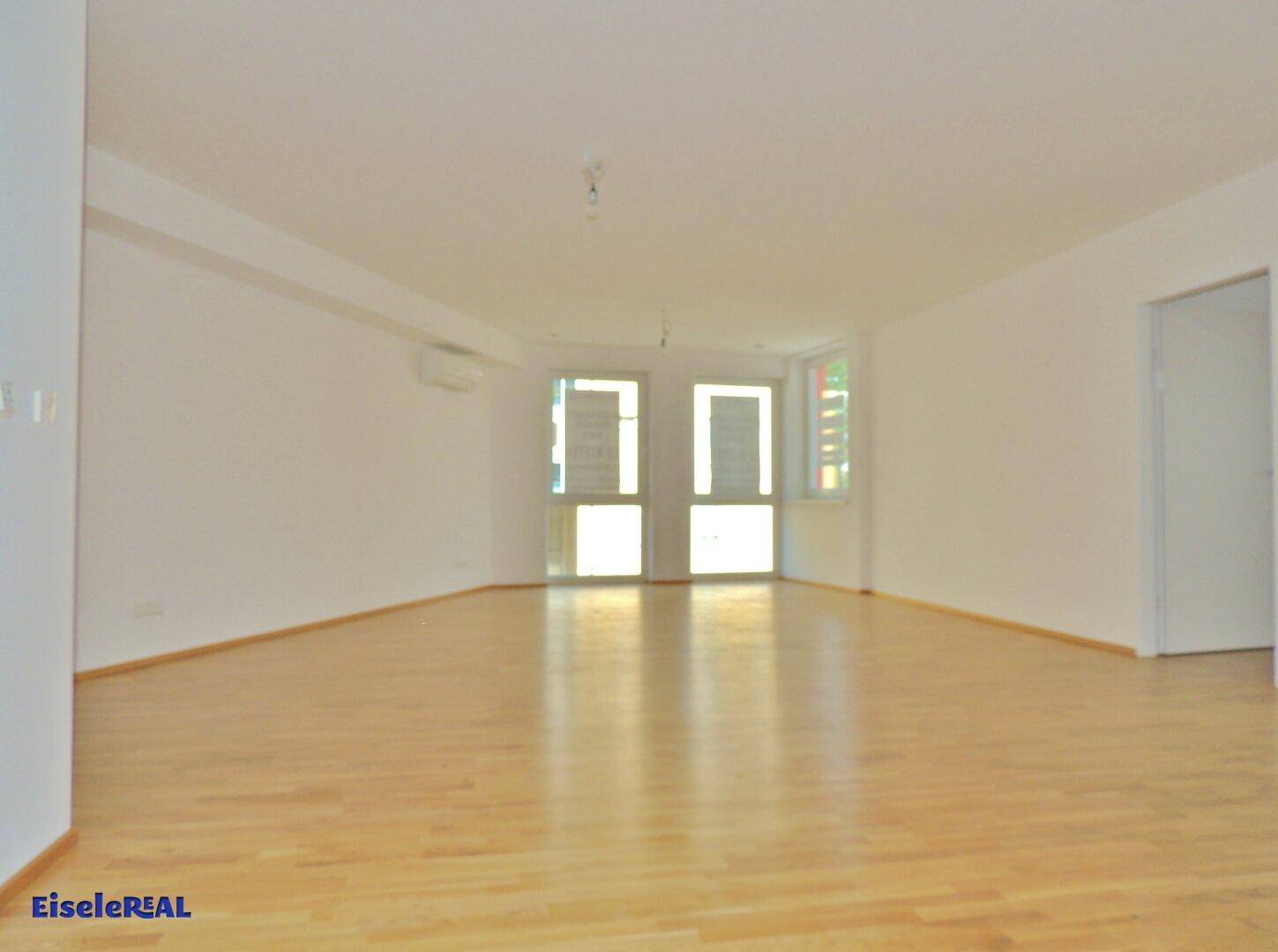 Heller, leerer Raum mit hellem Holzparkett und großen Fenstertüren, die viel Tageslicht hereinlassen und eine Klimaanlage zeigen.