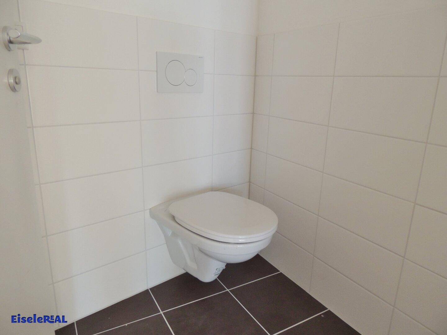 Modernes Gäste-WC mit wandhängender Toilette, weißen Fliesen an den Wänden und dunklen Bodenfliesen in sauberem Zustand.
