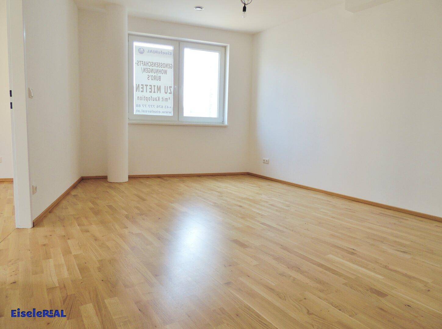 Heller, leerer Raum mit hellem Holzparkett, weißen Wänden und einem großen Fenster, das viel Tageslicht hereinlässt.