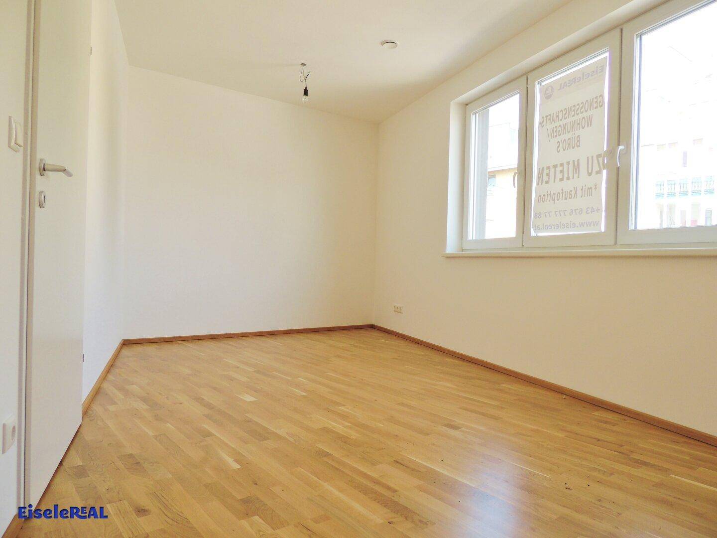 Geräumiger, leerer Raum mit hellem Holzparkett, weißen Wänden und einem großen Fenster, das viel Licht spendet und eine saubere Umgebung zeigt.