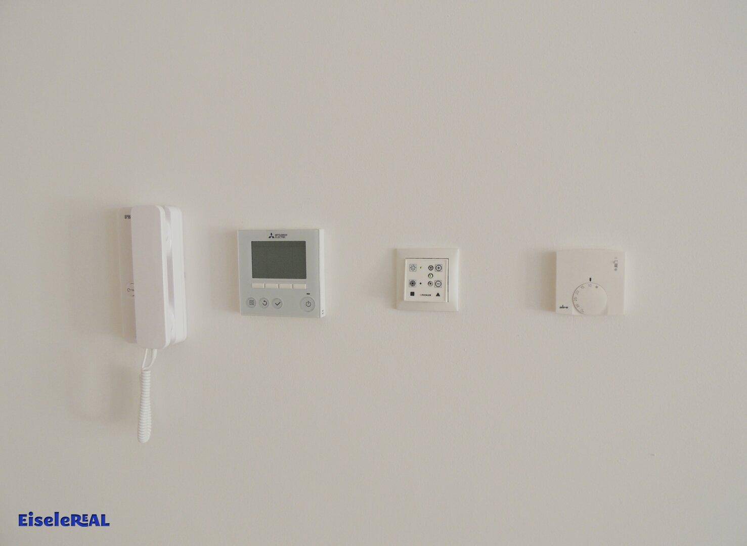 Verschiedene weiße Wandgeräte, darunter eine Gegensprechanlage und Thermostate, sind auf einer hellen Wand angebracht.