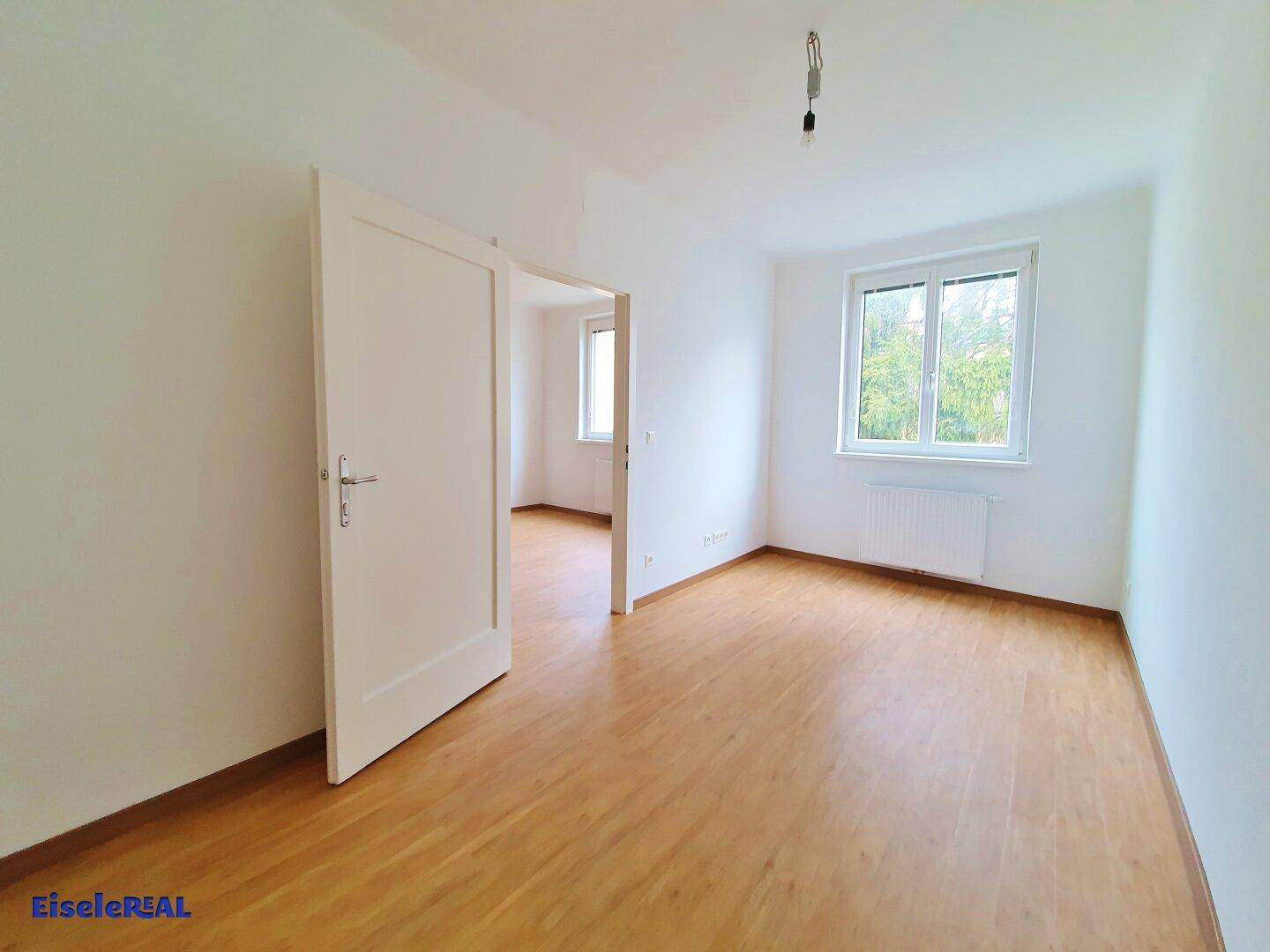 Geräumiges, helles Zimmer mit Holzboden und offener Tür zu einem weiteren Raum.