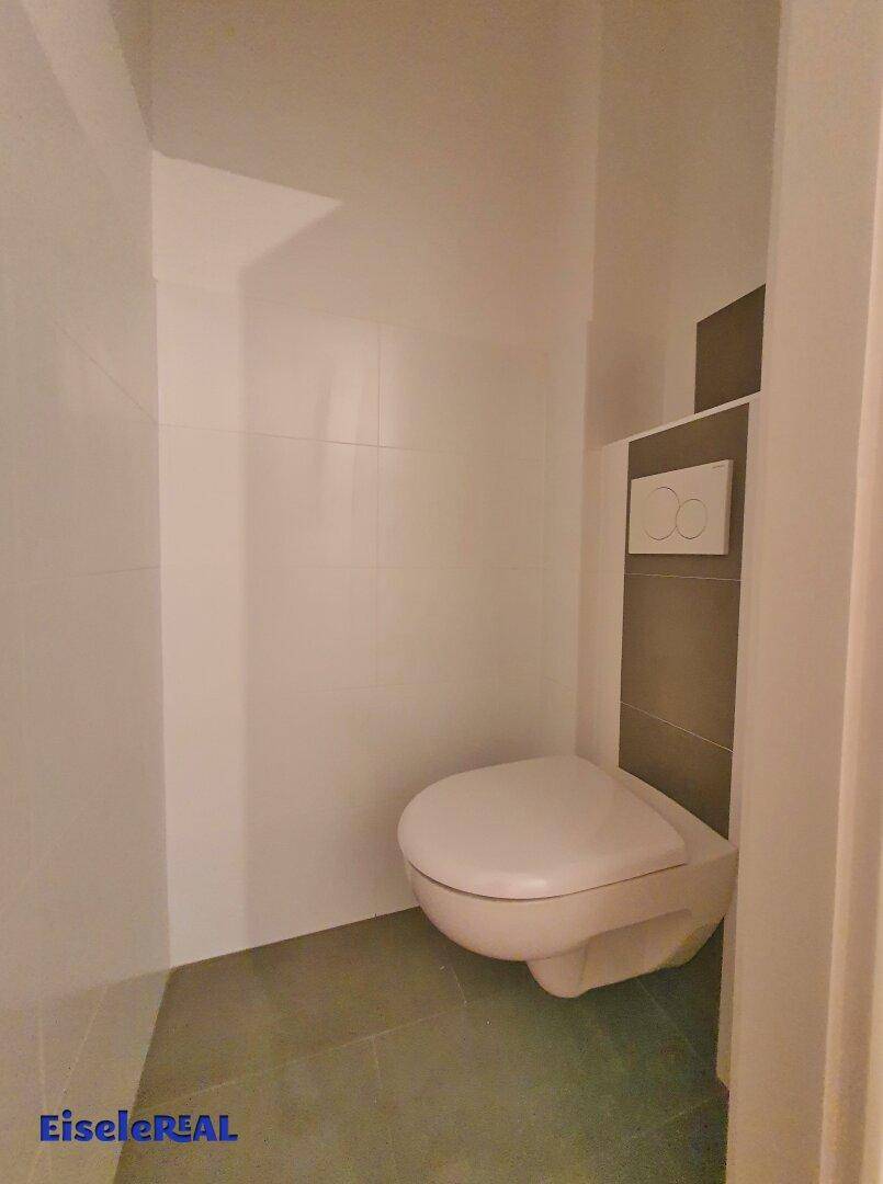 Separates WC mit wandhängender Toilette und modernen Fliesen in Weiß und Grau.