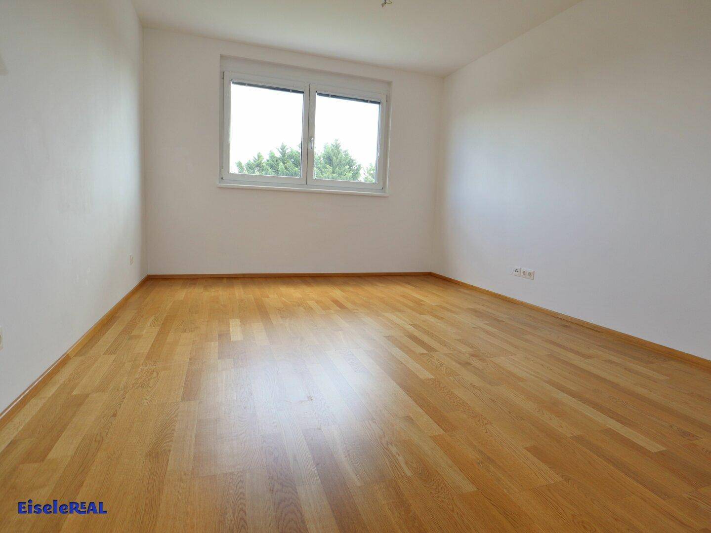 Geräumiges Zimmer mit Parkettboden und großem Fenster mit Blick ins Grüne.