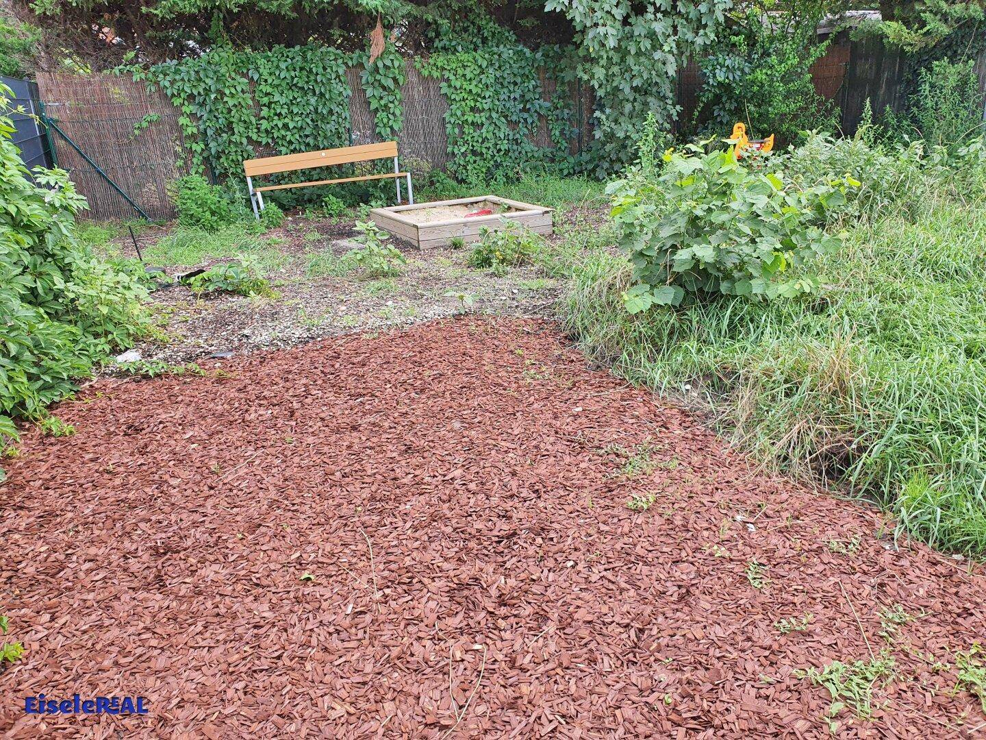 Gemeinschaftsgarten mit Sandkasten und Sitzbank, umgeben von Grünflächen.