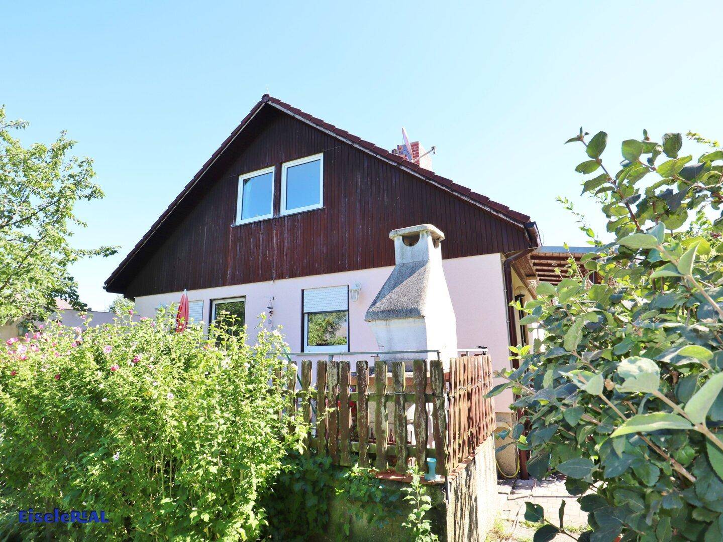 Seitenansicht des Hauses mit kleiner Holzterrasse und gemauertem Grill im Freien.