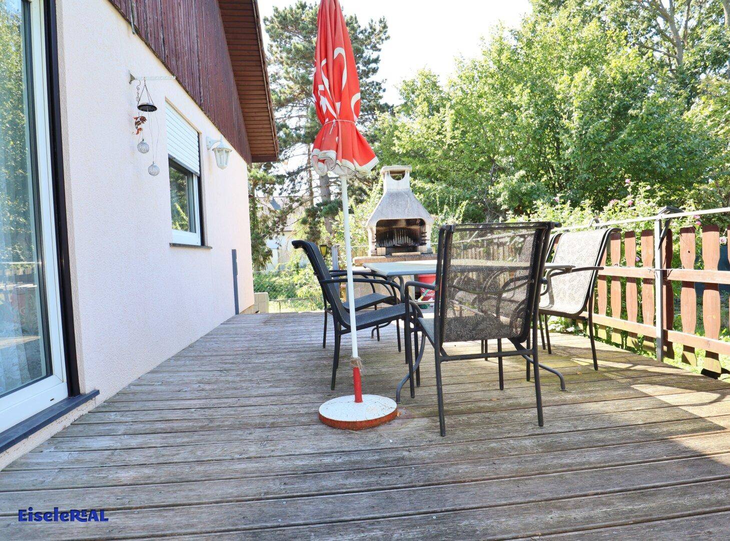 Holzterrasse mit Sonnenschirm und gemauertem Grill im Hintergrund, umgeben von Bäumen.