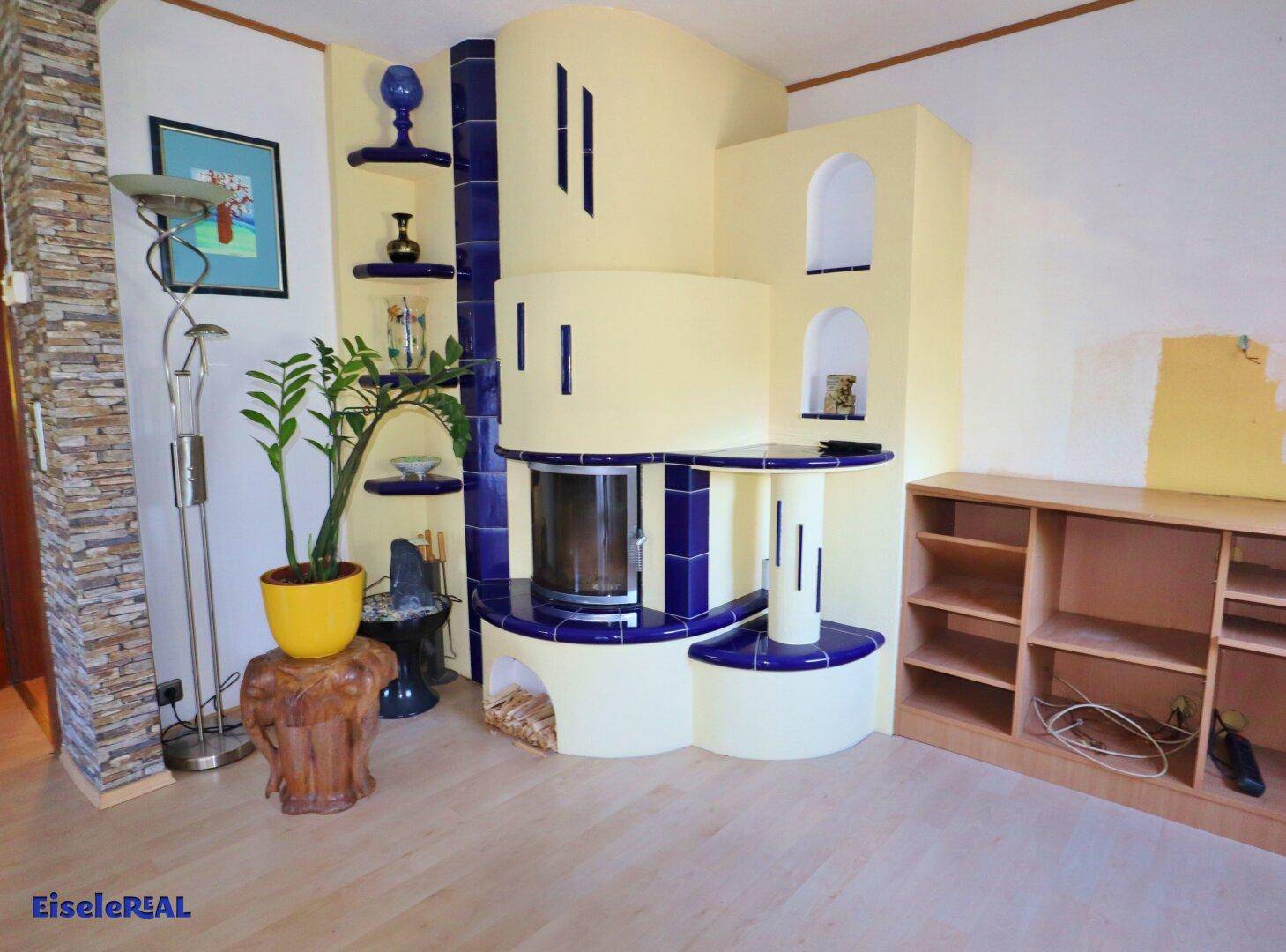 Helles Wohnzimmer mit einem auffälligen blau-gelben Kachelofen und hellem Laminatboden.