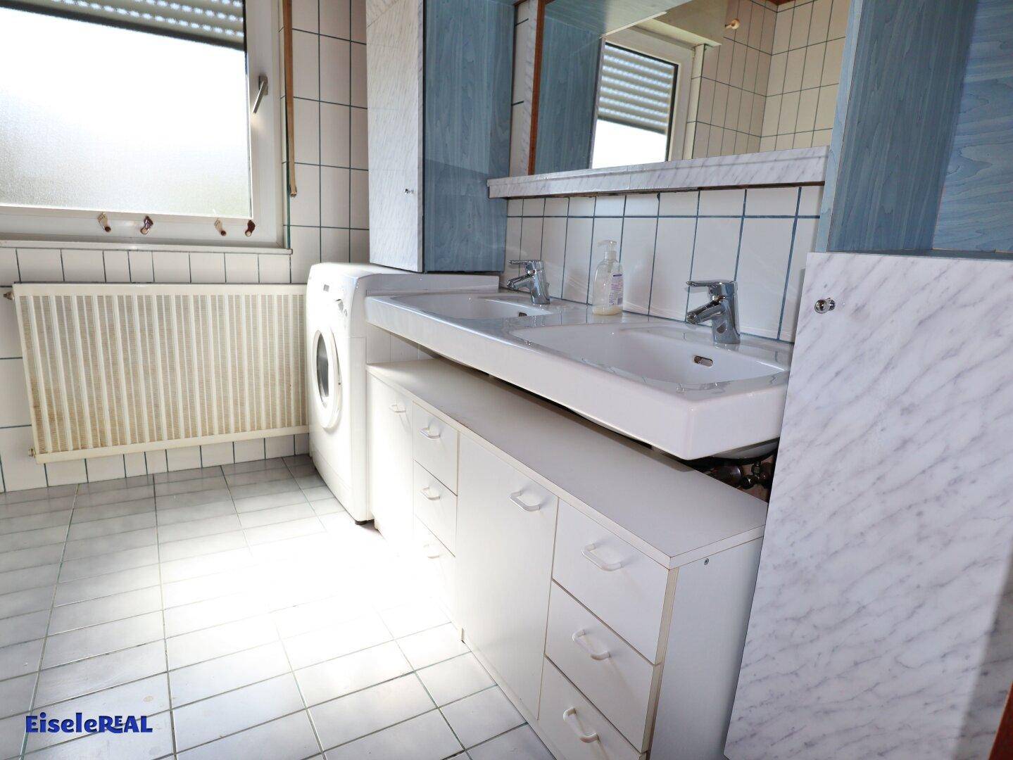 Helles Badezimmer mit Doppelwaschbecken, Waschmaschine und weißen Fliesen.