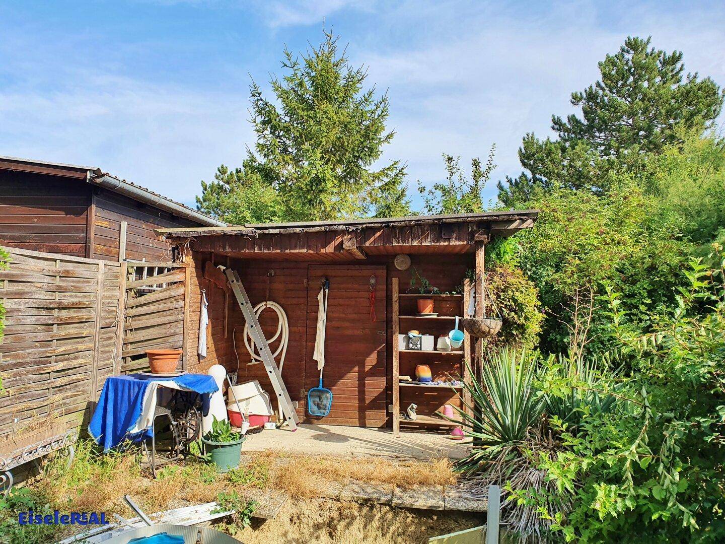 Rustikaler Holzschuppen im Garten mit Regalen und Gartengeräten, umgeben von Pflanzen.