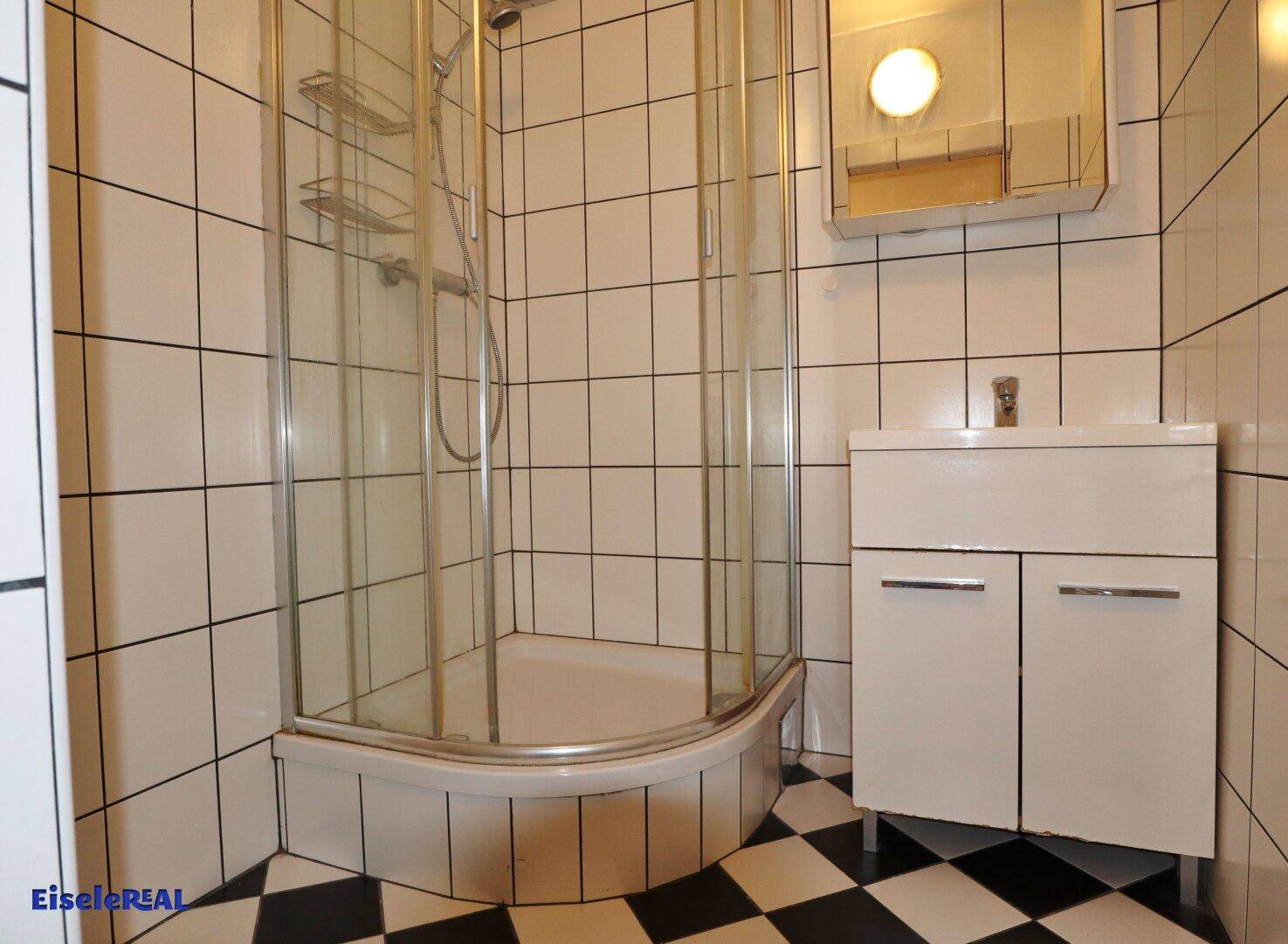 Badezimmer mit Duschkabine, Waschbeckenunterschrank und schwarz-weiß kariertem Fliesenboden.