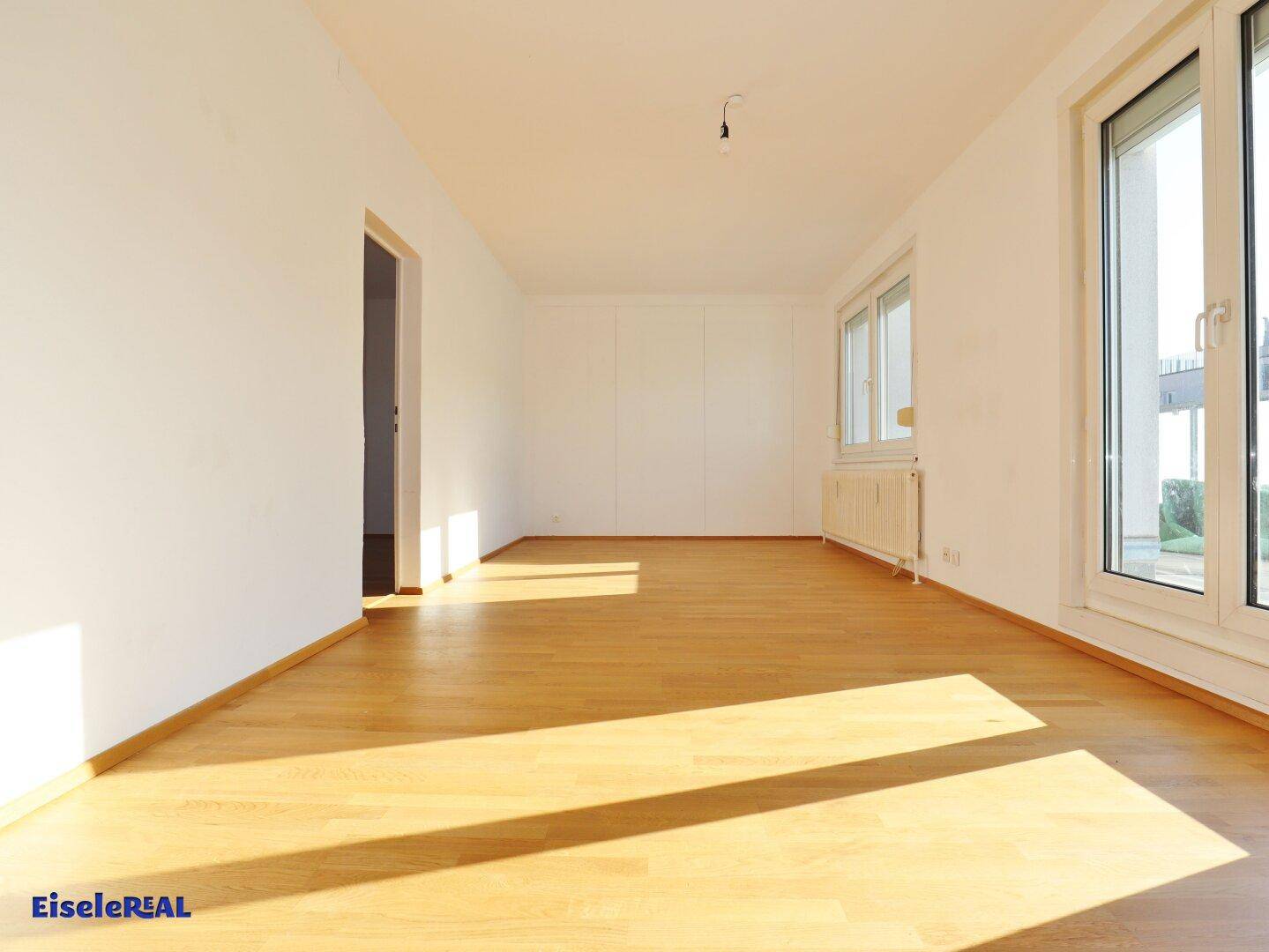 Helles, leeres Wohnzimmer mit hellem Parkettboden und großen Fenstern, die viel Sonnenlicht hereinlassen.