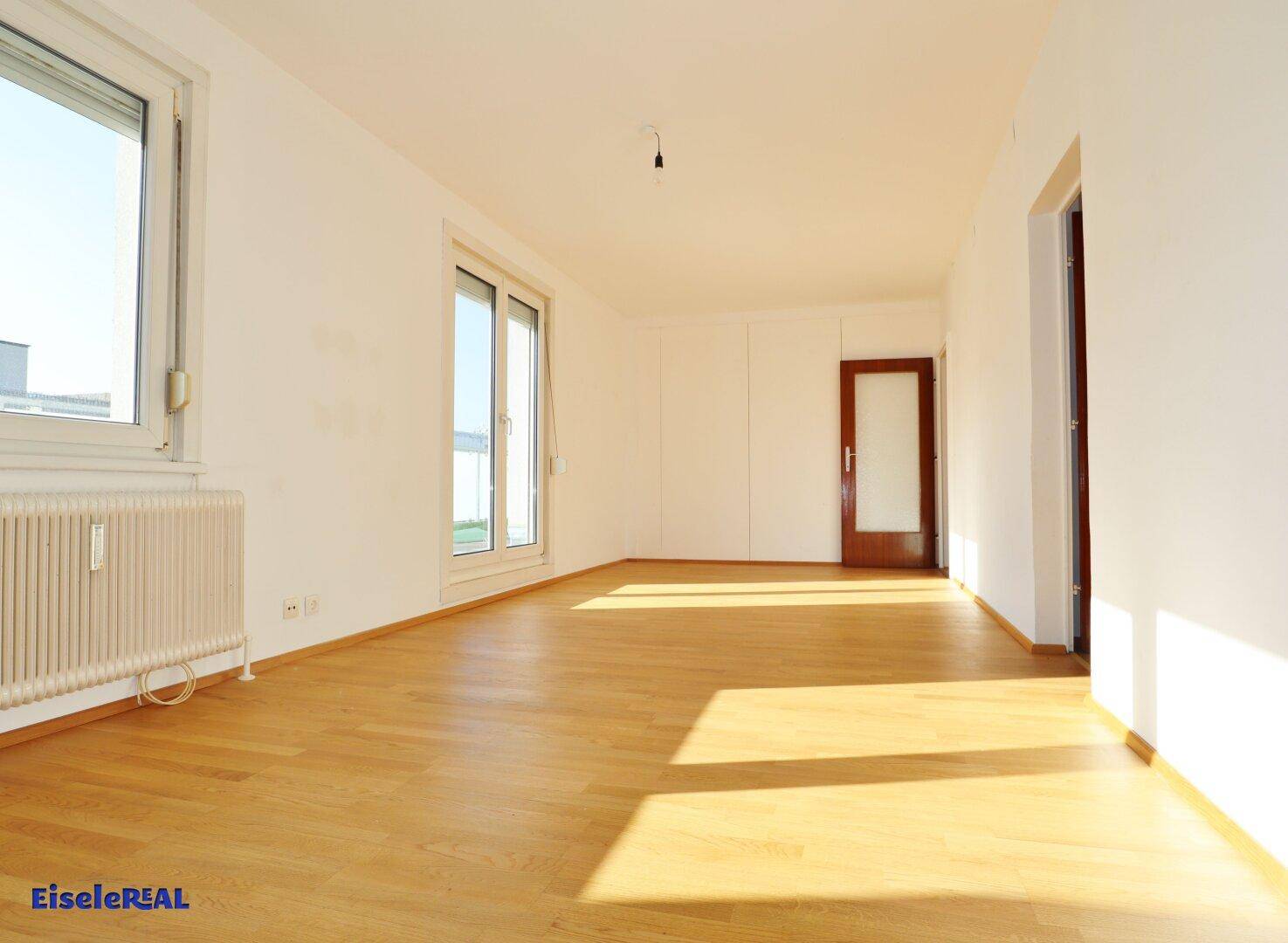 Geräumiges, leeres Wohnzimmer mit Holzboden und Zugang zu einem Außenbereich durch eine Glastür.