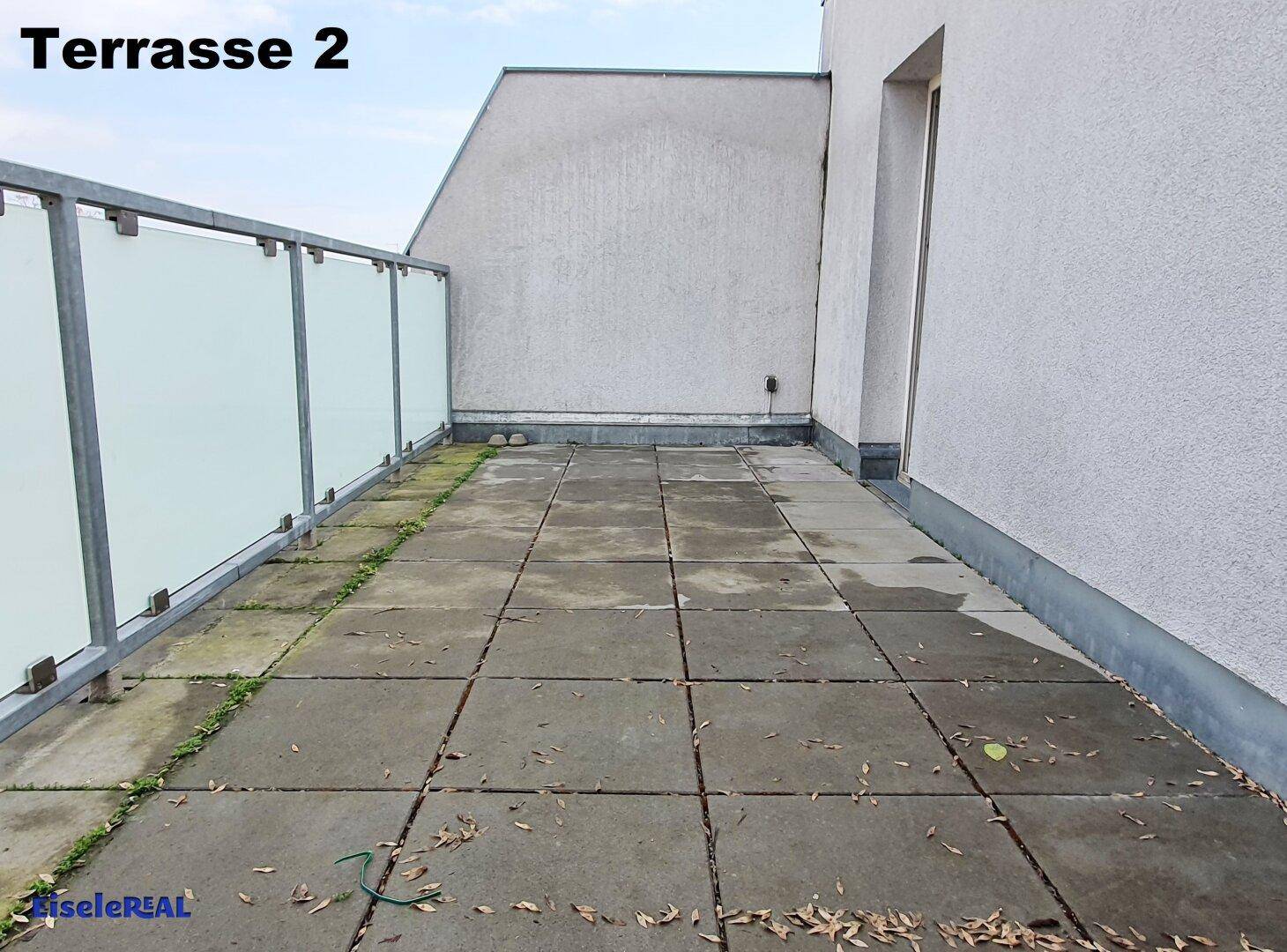Weitläufige Dachterrasse mit Fliesenboden und Glasgeländer, umgeben von Gebäuden.