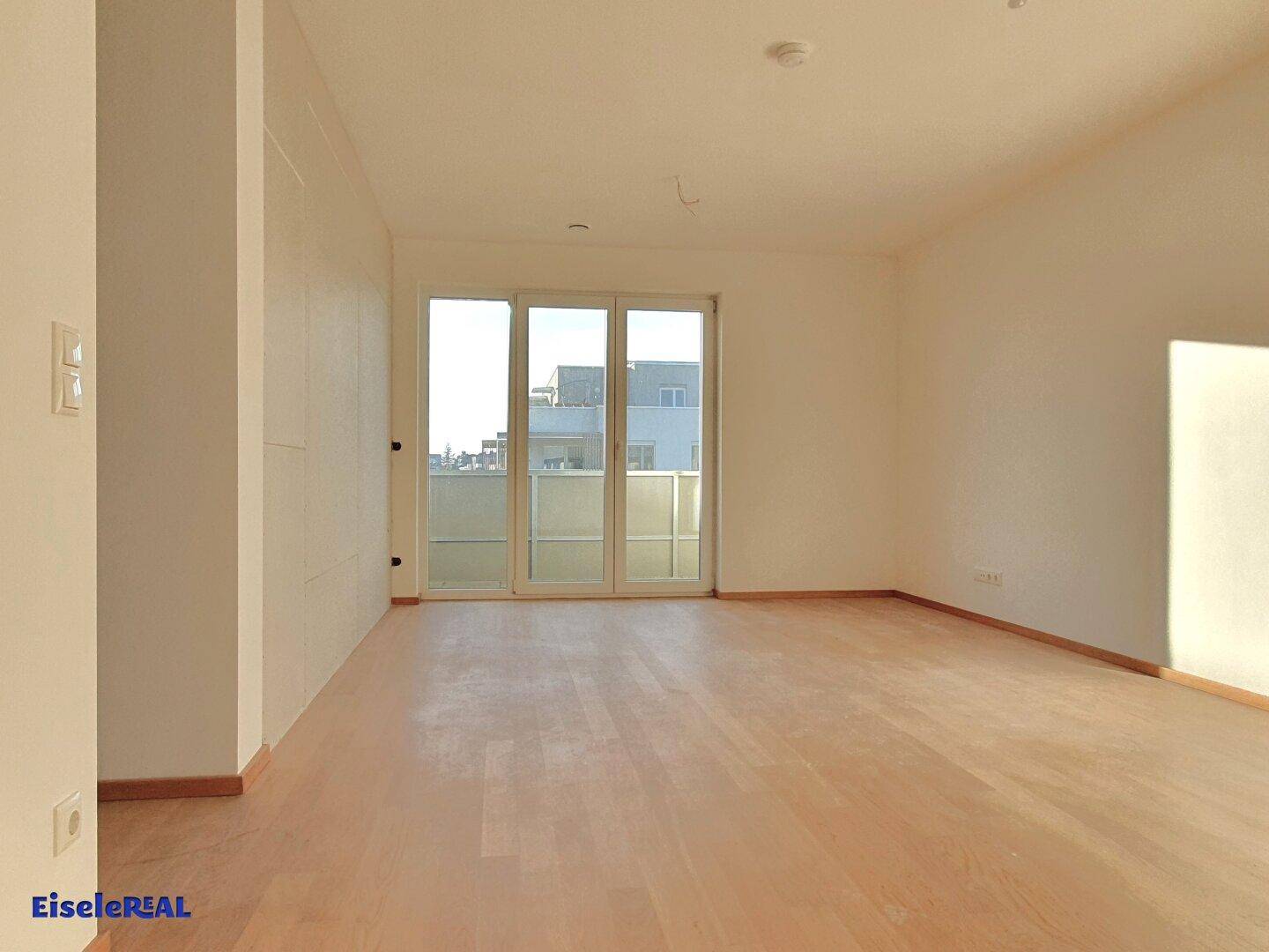 Heller, leerer Wohnraum mit Holzboden und großem Balkonzugang, der viel Tageslicht hereinlässt.