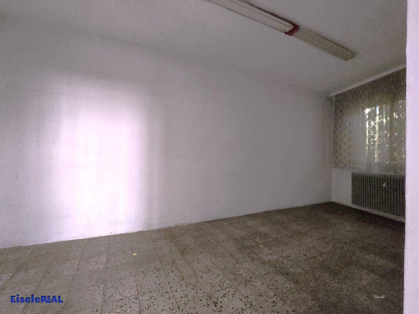 Kleiner, leerer Raum mit Terrazzoboden, Heizkörper und einem Fenster mit Vorhang.